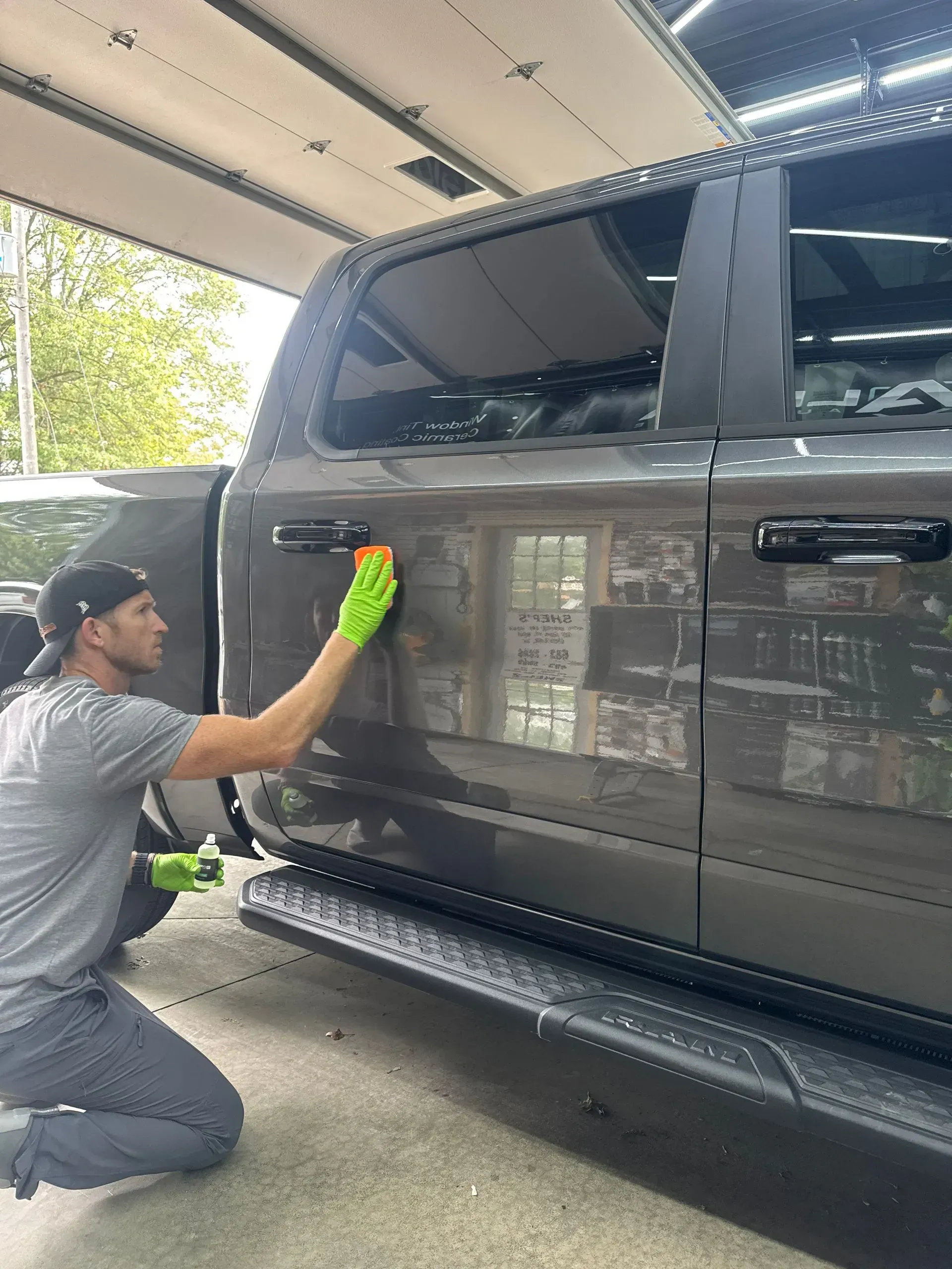 Person kneeling, cleaning the side of a black pickup truck in a covered driveway.