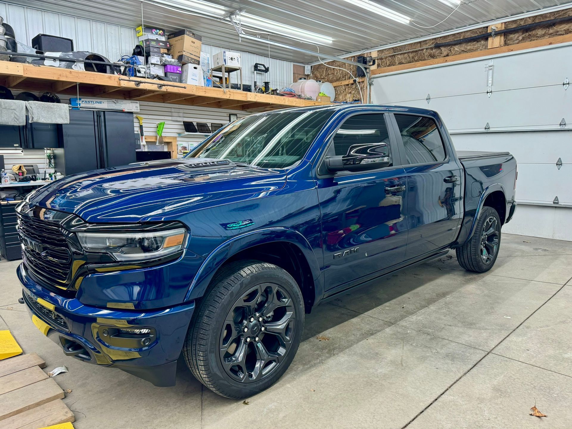 Blue pickup truck in a garage workshop, angled front view with black wheels and yellow tow hooks.