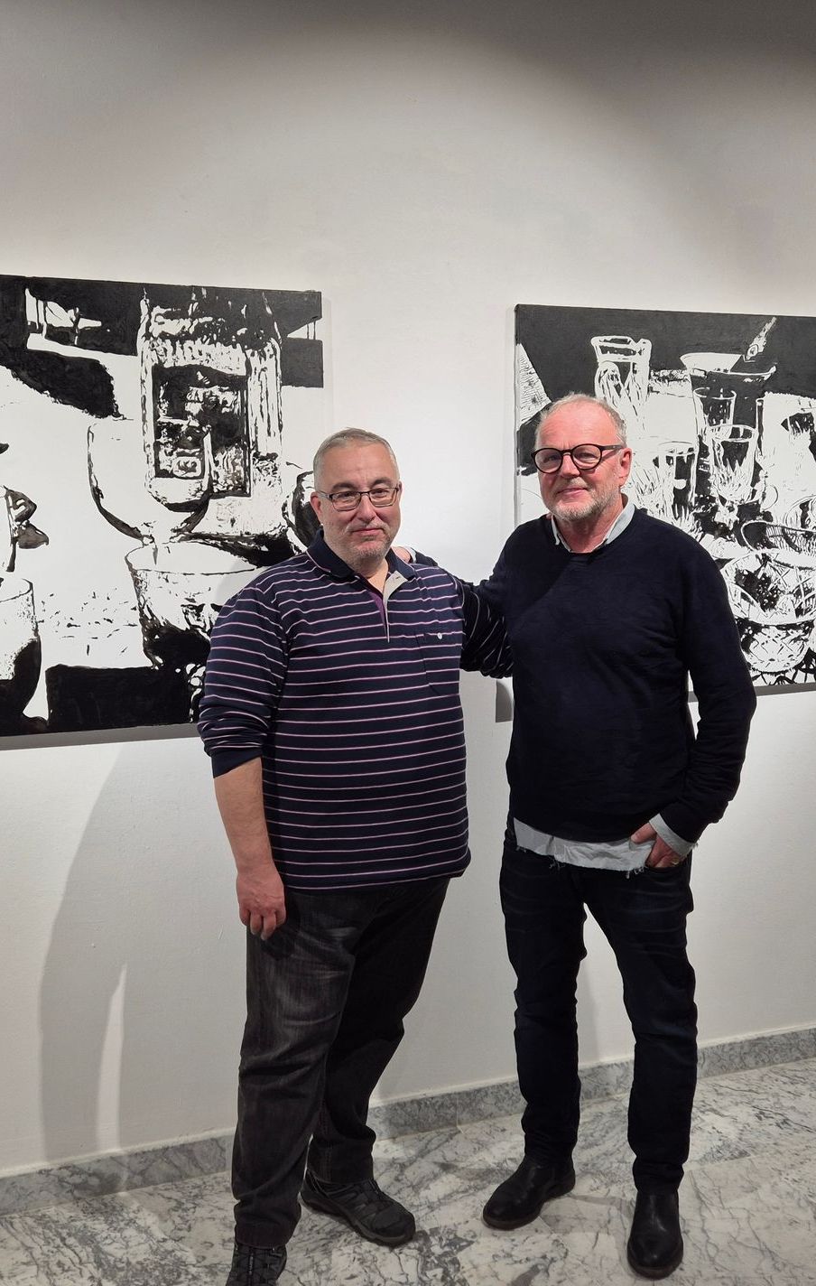 Two men pose in an art gallery next to black and white abstract art.