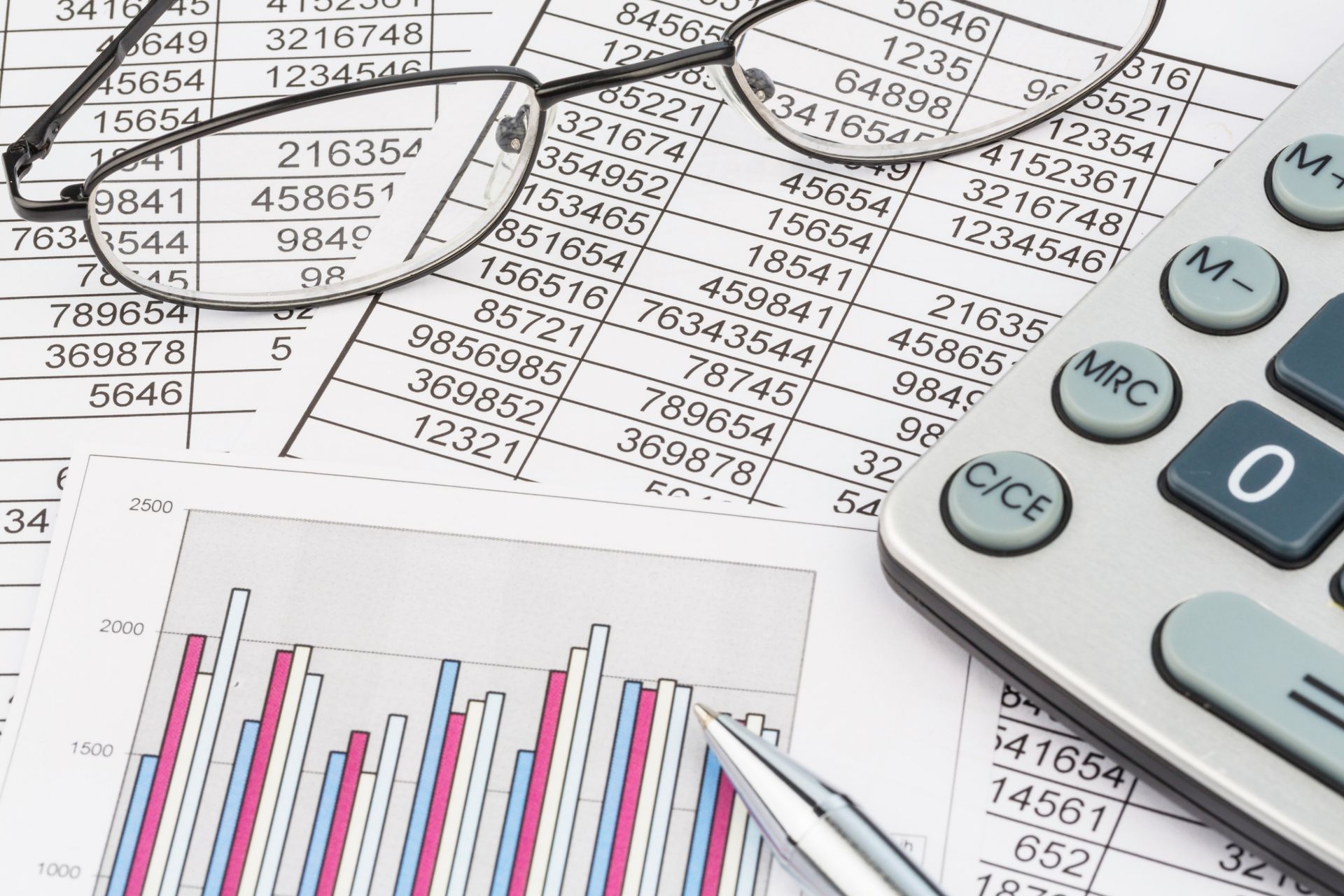 Calculator, eyeglasses, charts, and financial documents on a desk with spreadsheets and numbers.