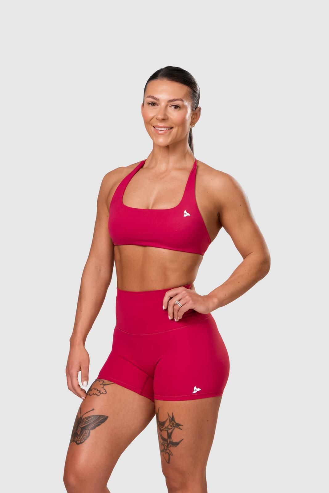 Woman lifting dumbbells, arm flexed, in black top and white shorts. Red background.