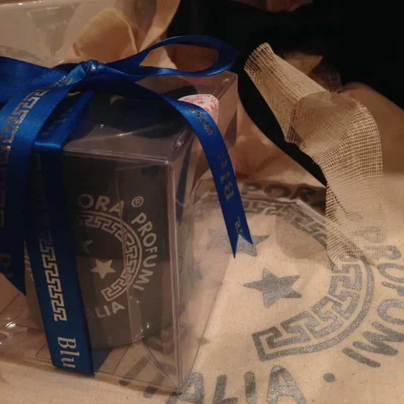 Scatola regalo avvolta in plastica trasparente con nastro blu, accanto a un sacchetto di stoffa color beige 