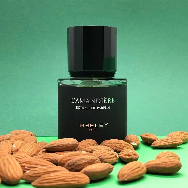 Flacone di profumo L'Amandière con tappo nero, circondato da mandorle su sfondo verde.