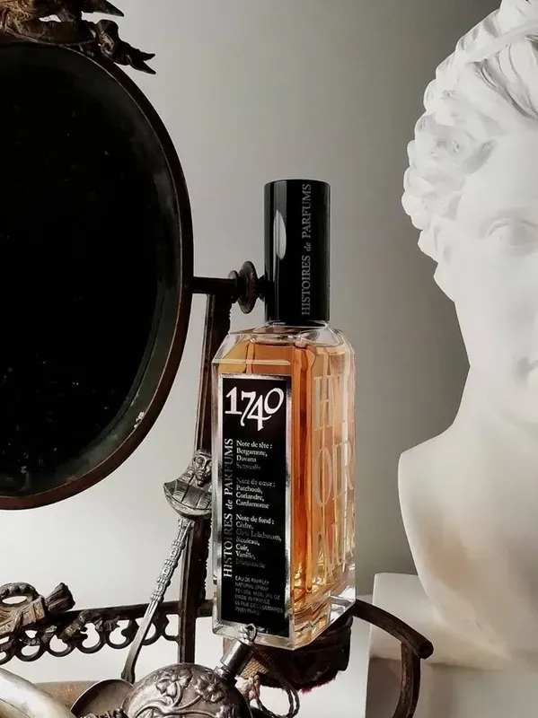 Flacone di profumo con etichetta nera su una toeletta accanto a uno specchio rotondo e a una scultura a busto bianca.