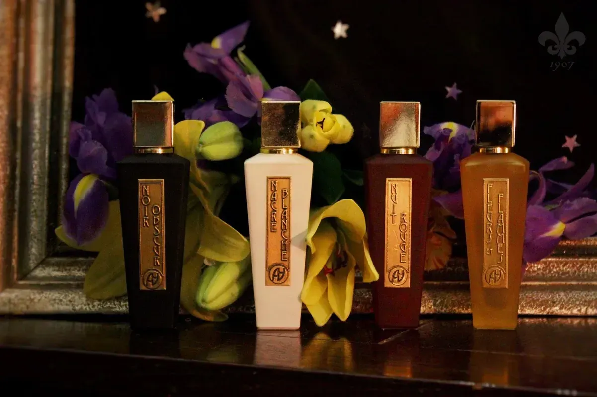Cinque flaconi di profumo con tappi dorati disposti su una superficie scura con fiori viola e gialli sullo sfondo.