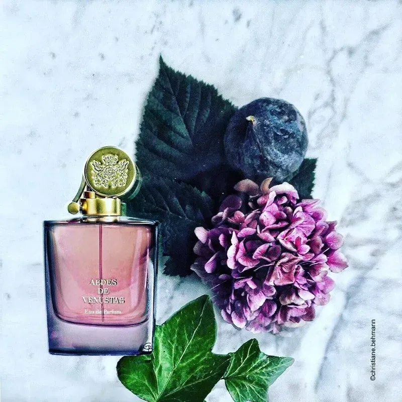 Flacone di profumo rosa con tappo dorato accanto a fiori viola e foglie verde scuro su uno sfondo di marmo chiaro.