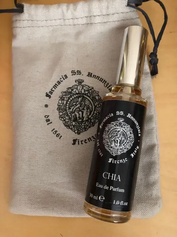Flacone di profumo trasparente su una pochette beige con coulisse, etichetta nera e tappo dorato.