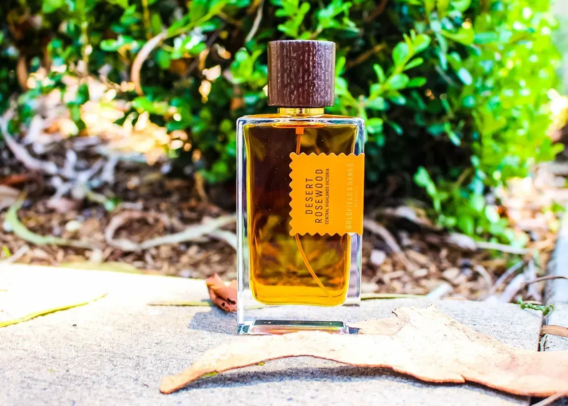 Flacone di profumo color ambra con tappo di sughero su un tavolo all'aperto illuminato dal sole, su uno sfondo verde sfocato.