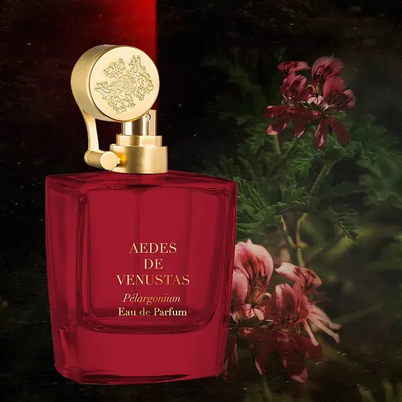 Flacone di profumo rosso con tappo dorato ed emblema floreale su sfondo scuro.