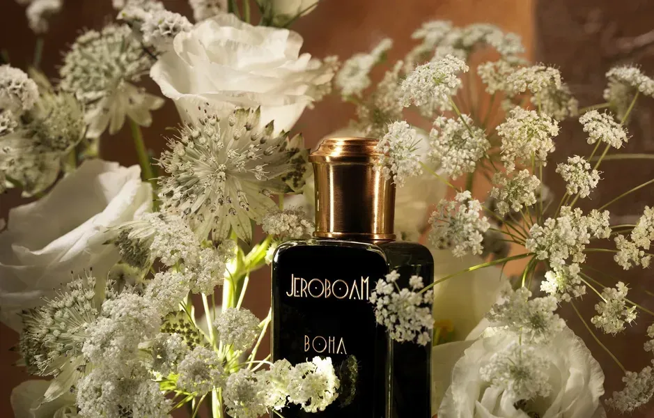Flacone di profumo nero con tappo dorato tra fiori bianchi e vegetazione.