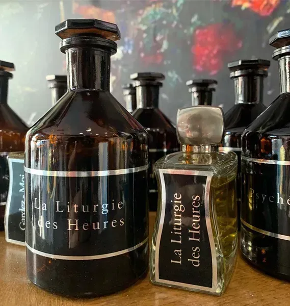 Bottiglie di profumo in vetro scuro etichettate “La Liturgie des Heures” su una superficie riflettente.