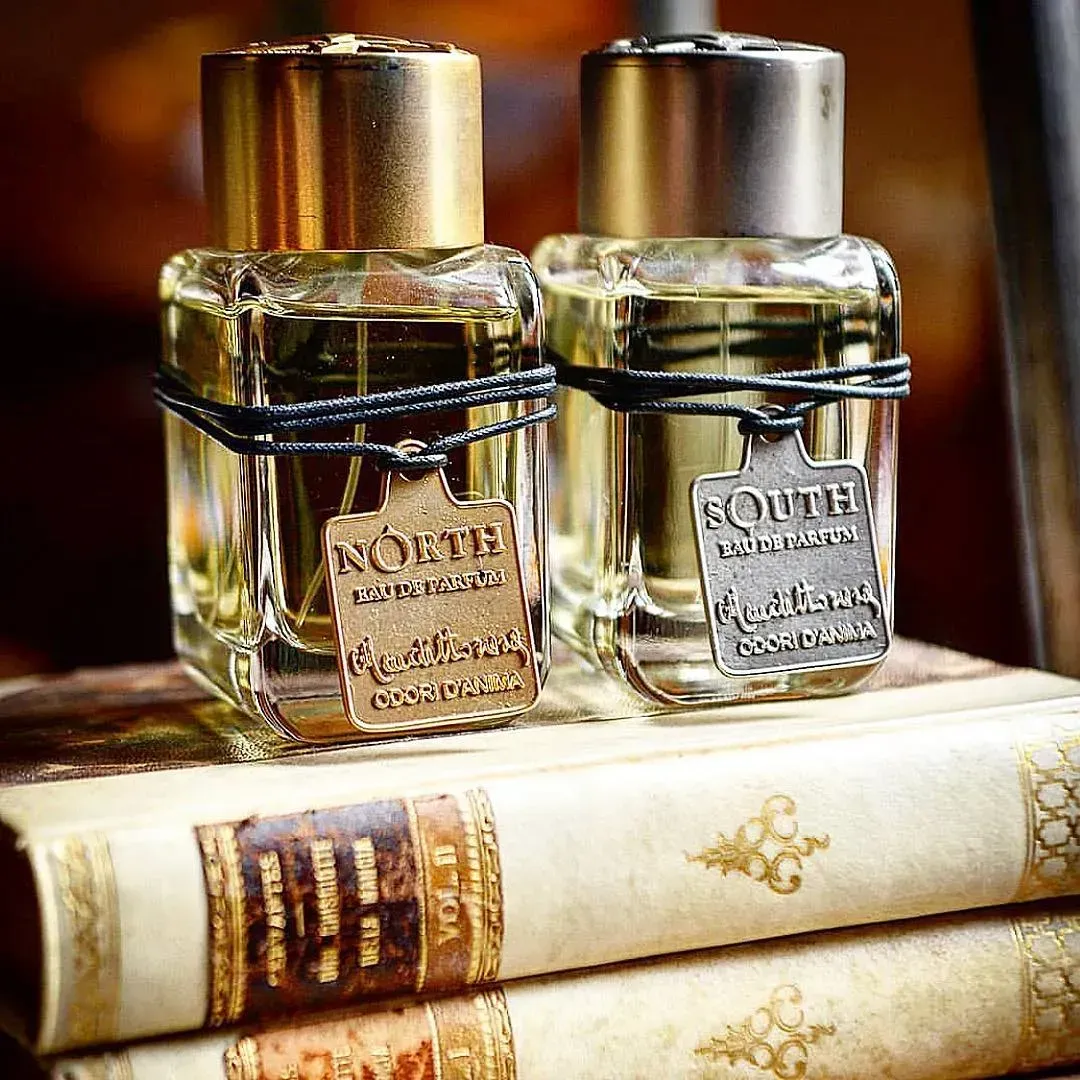Due piccole boccette di profumo in vetro con tappi dorati, appoggiate su una pila di libri vintage.