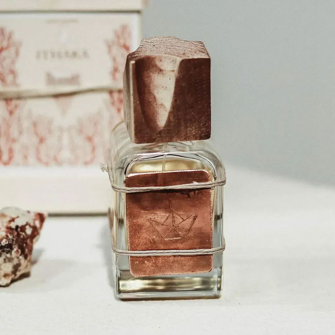 Flacone di profumo trasparente con tappo color rame su una superficie bianca, contro un delicato sfondo floreale.