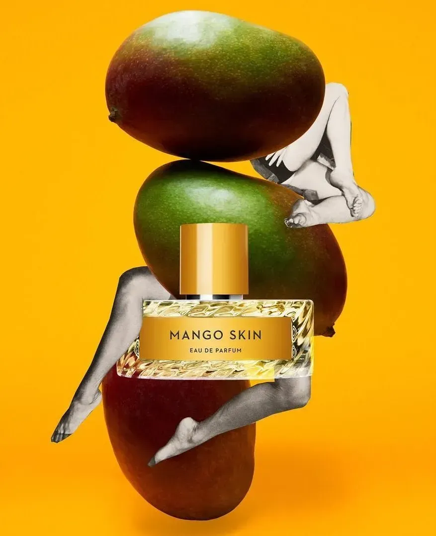 Flacone di profumo Mango Skin su sfondo arancione brillante con manghi impilati e dettagli argentati.