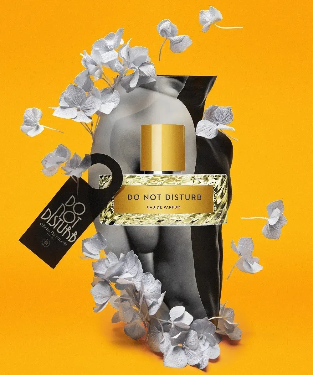 Flacone di profumo nero e oro circondato da fiori bianchi su uno sfondo arancione brillante.