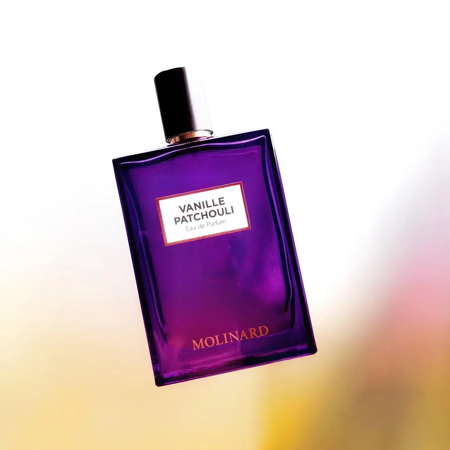 Flacone viola di profumo alla vaniglia e patchouli su sfondo bianco con delicate sfumature rosa e gialle.