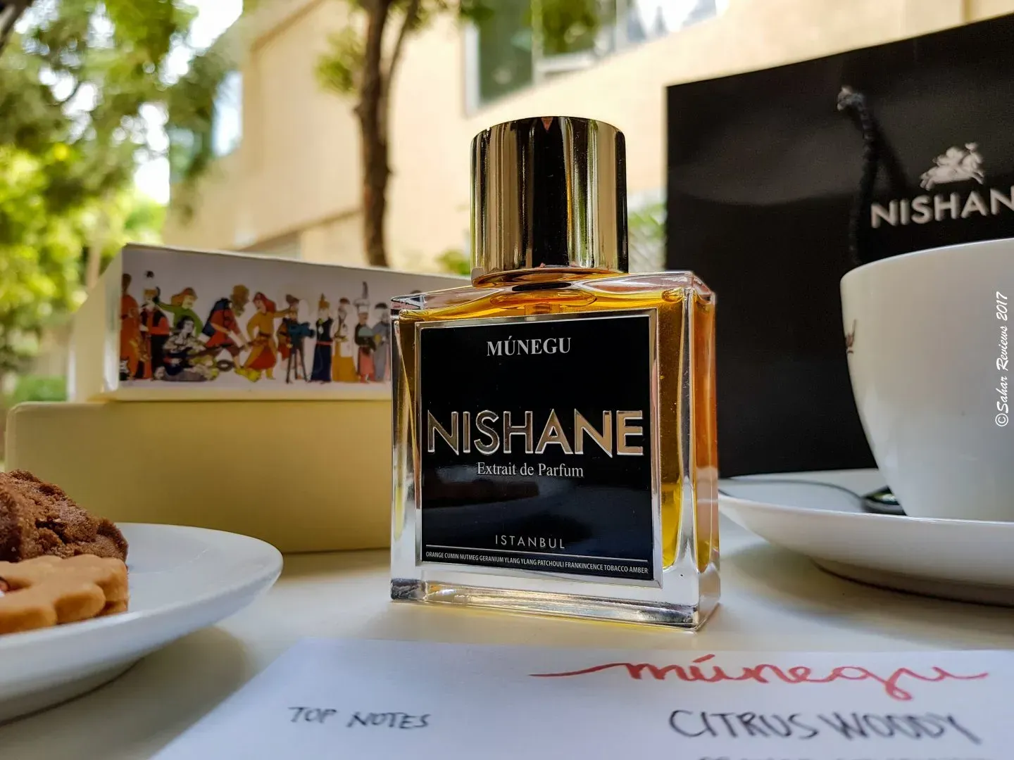 Una boccetta di profumo Nishane su un tavolo con piattini da dessert, una tazza e la calda luce del sole sullo sfondo.
