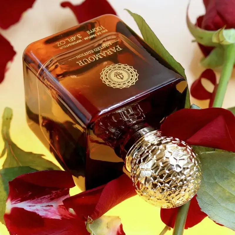 Flacone di profumo color ambra con tappo dorato, adagiato su petali di rosa rossa e foglie verdi.