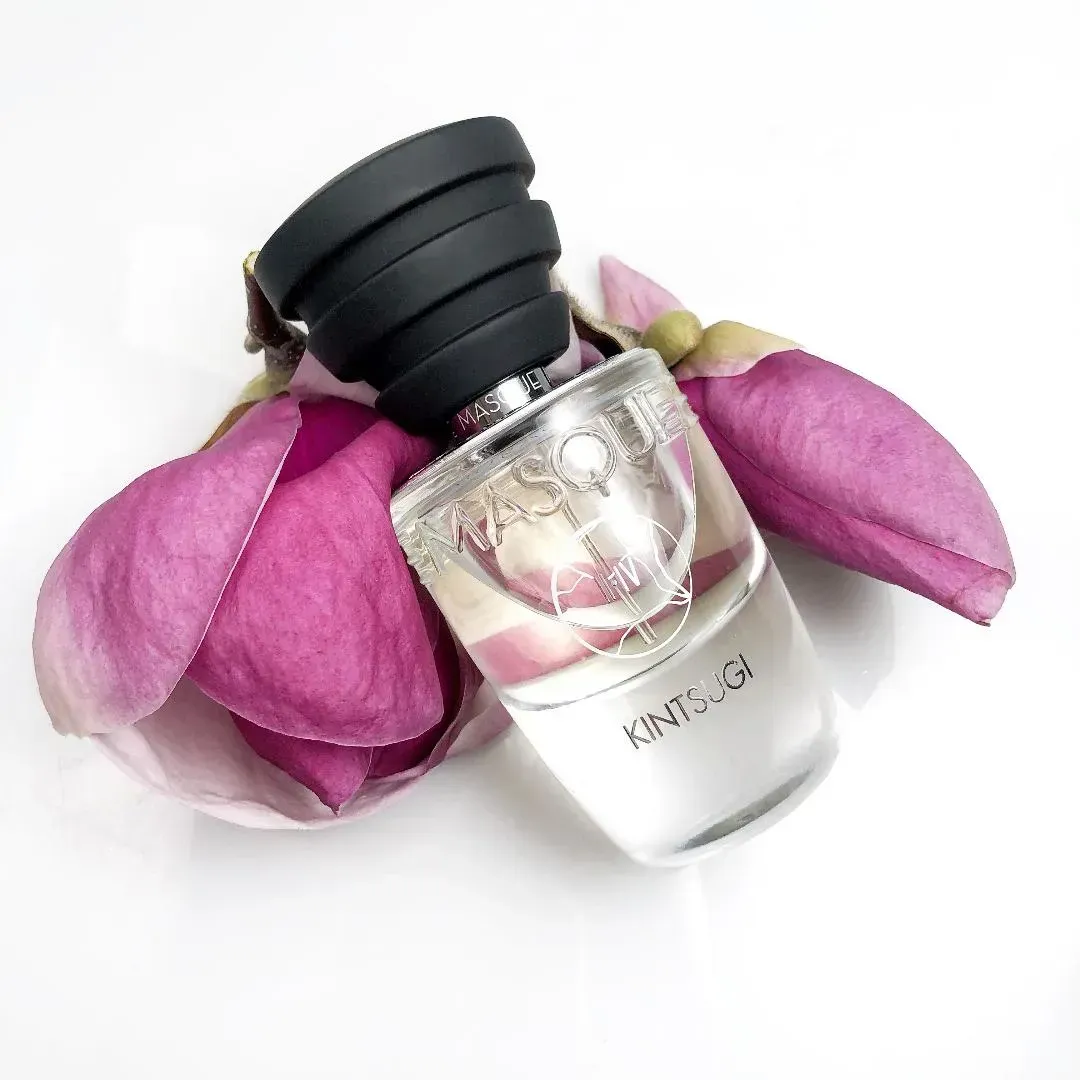 Flacone di profumo trasparente con tappo nero accanto a petali di fiori rosa su sfondo bianco.