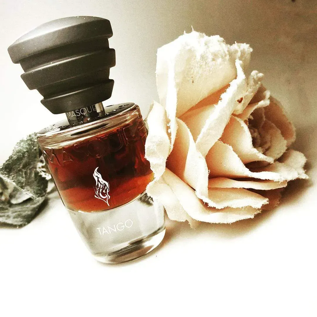 Flacone di profumo con tappo nero accanto a un fiore beige su sfondo bianco.