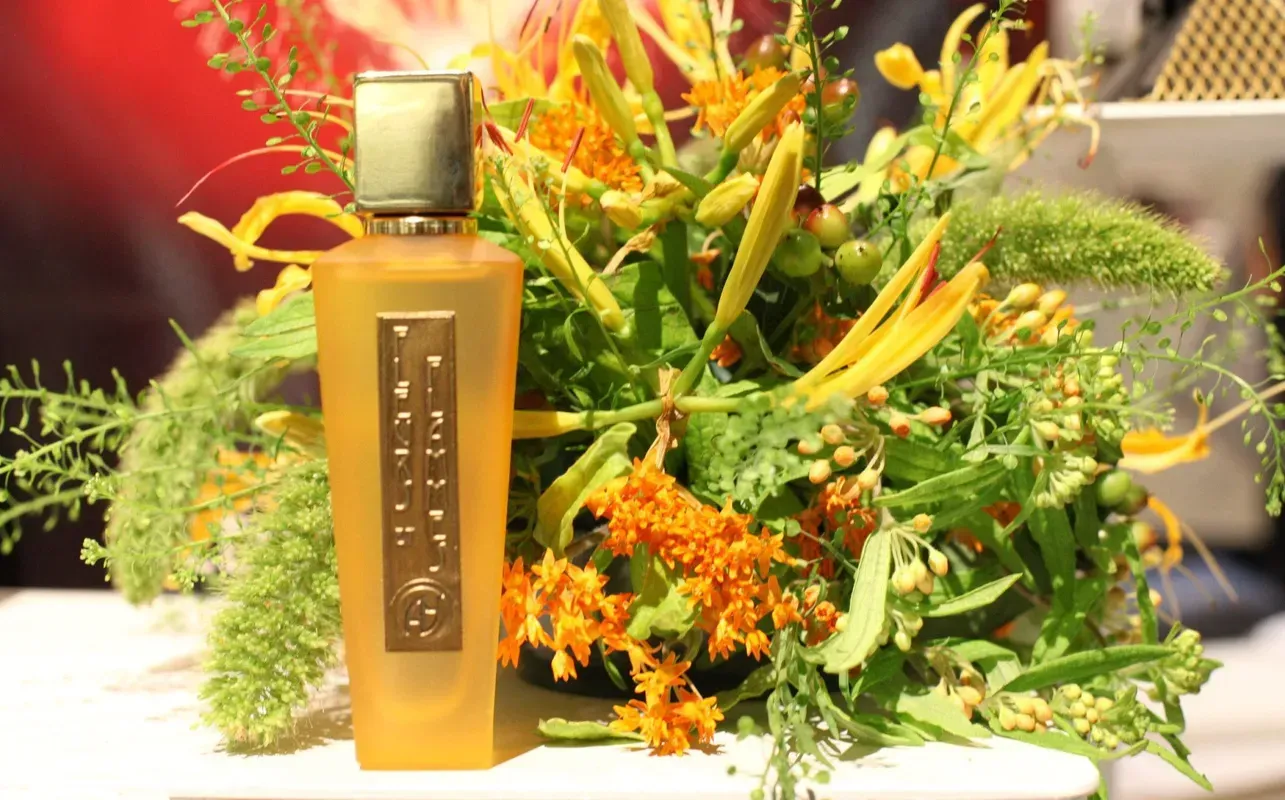 Flacone di profumo color ambra con tappo dorato, sullo sfondo di una composizione floreale giallo-verde brillante.