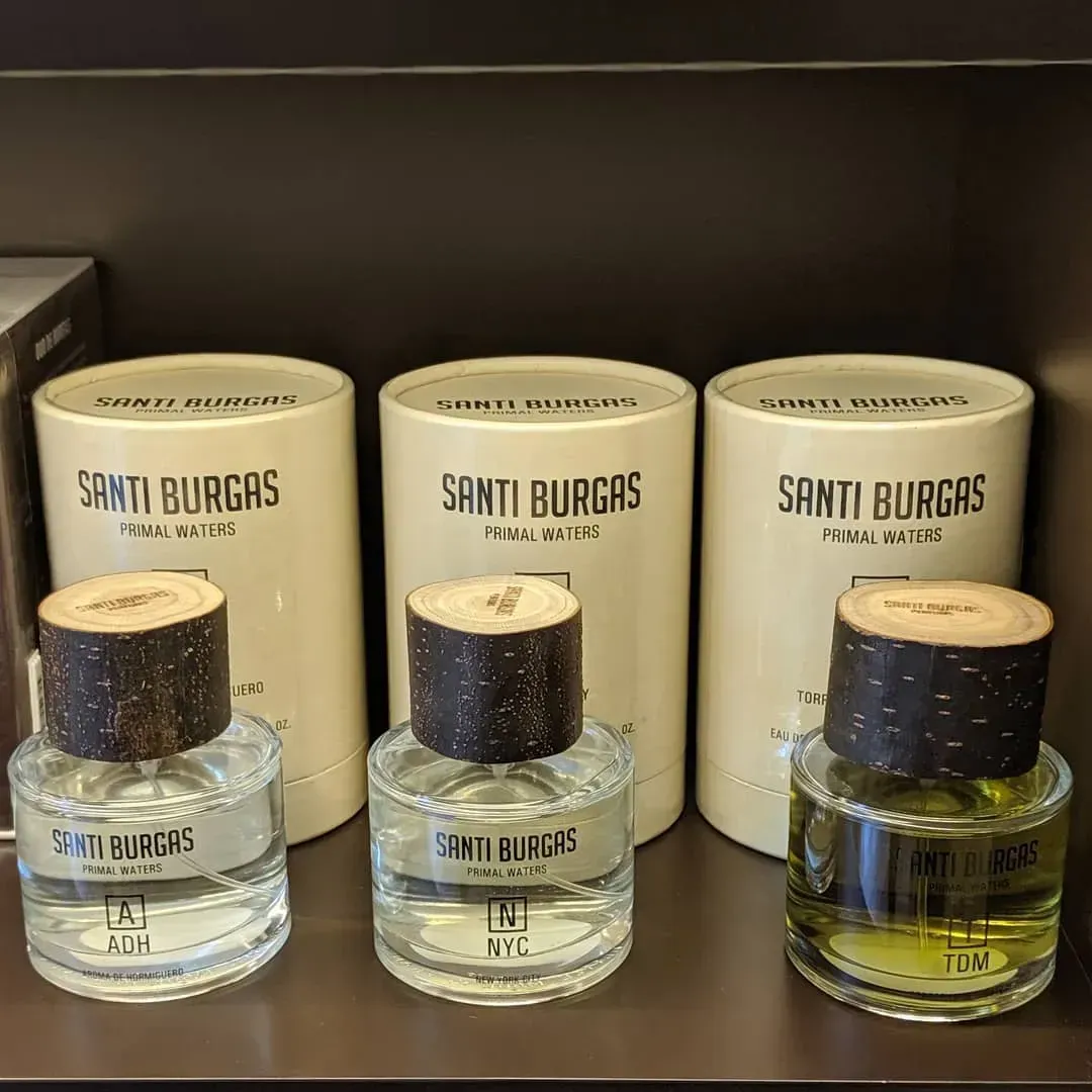 Tre flaconi di profumo SANTI BURGAS con tappo di sughero esposti davanti a scatole abbinate su uno scaffale.