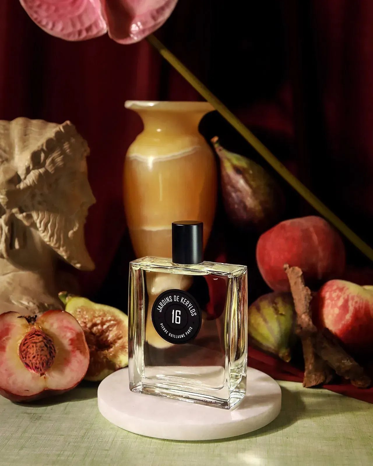 Flacone di profumo su un supporto bianco tra fichi, vaso e conchiglie, sullo sfondo di un drappeggio rosso scuro.
