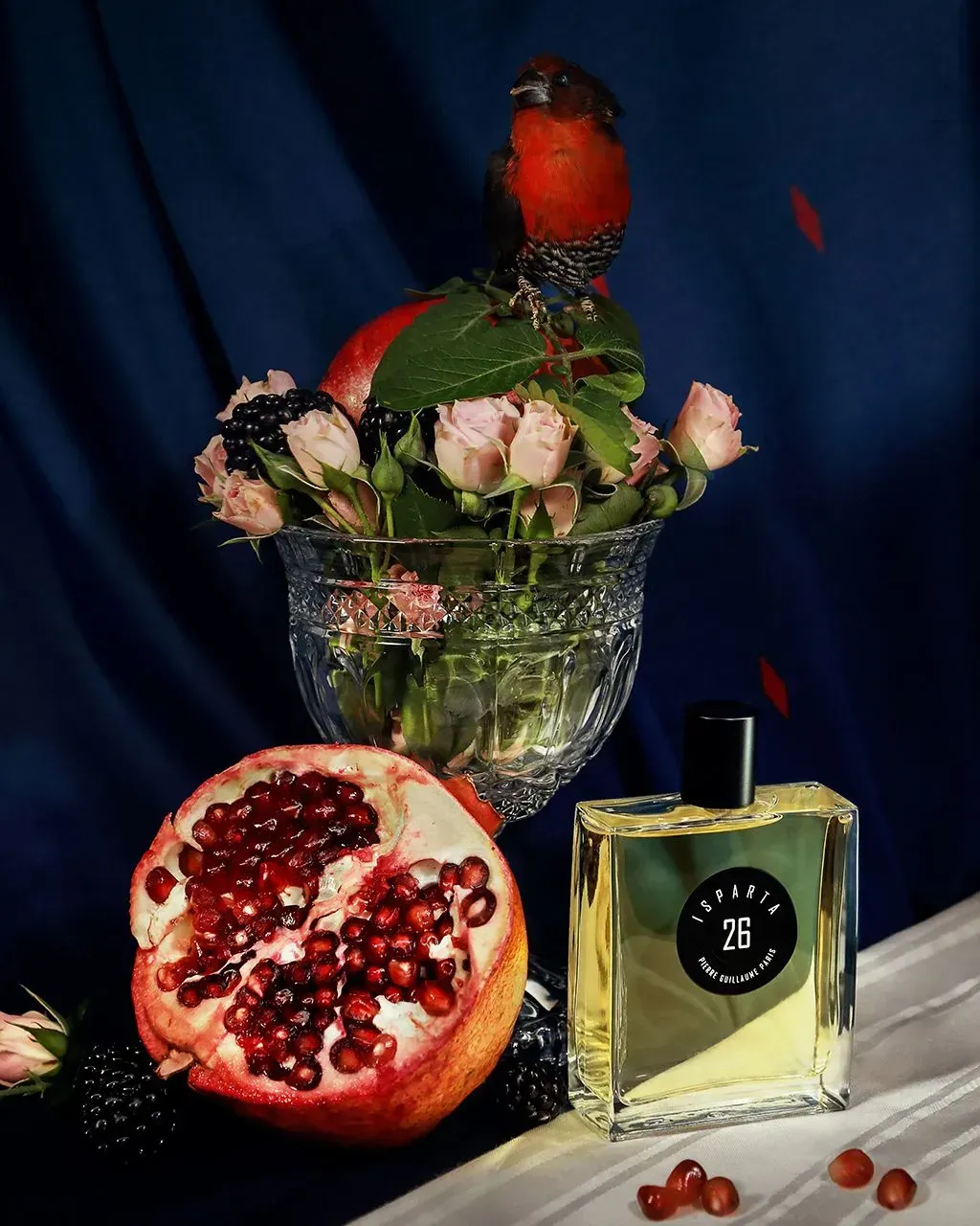Natura morta con un melograno rosso, fiori in un vaso, una boccetta di profumo e una statuetta di uccello su un telo scuro.