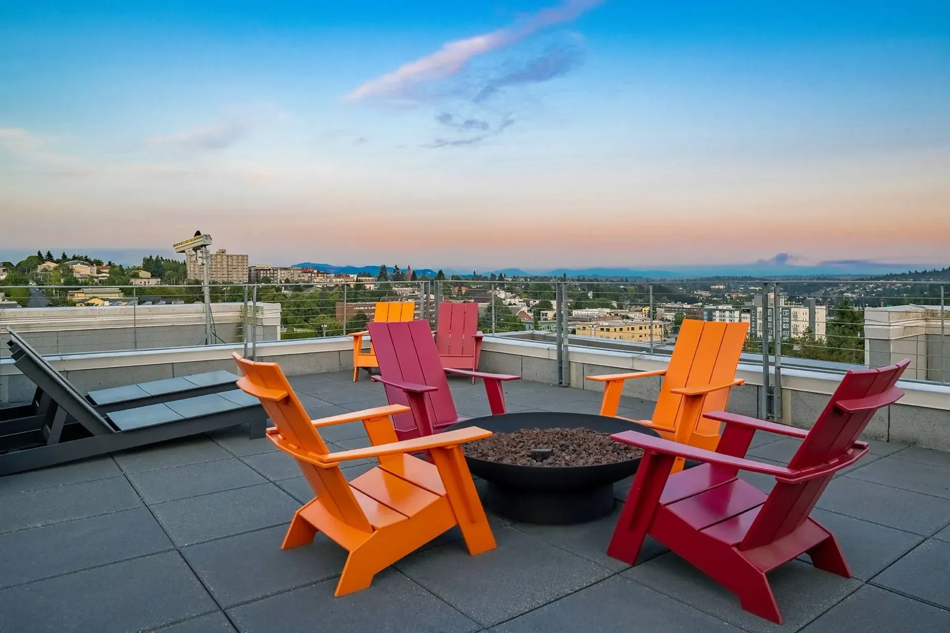 Rooftop patio with colorful Adirondack chairs around a fire pit, overlooking a city skyline at sunset.