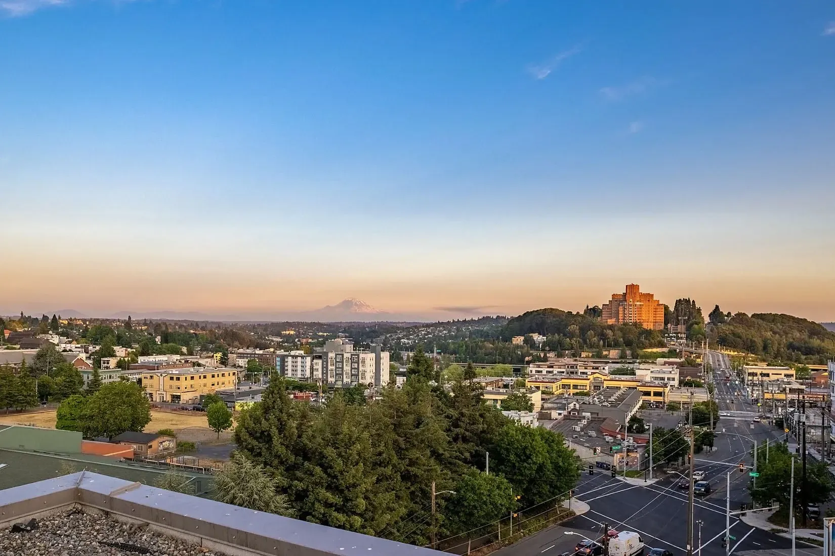 Panoramic view of a city skyline with mountains in the distance at sunset, showcasing surrounding buildings and roads.