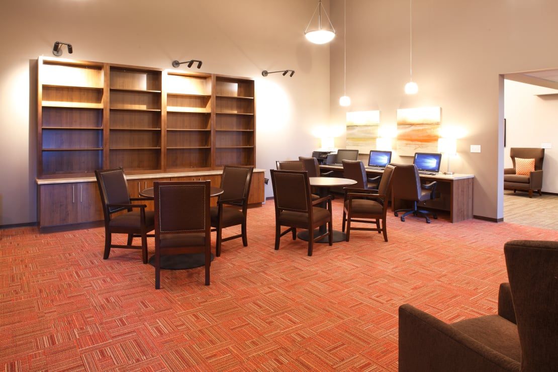 Interior of a modern community lounge with tables, chairs, computers, and tall bookshelves.
