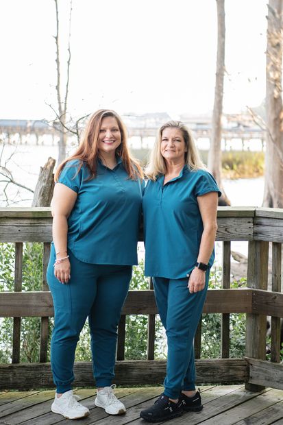Two women in teal scrubs standing outdoors on a wooden deck, smiling at the camera.