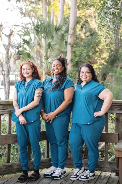 Three people in teal shirts and pants standing outdoors by a wooden fence, smiling at the camera.