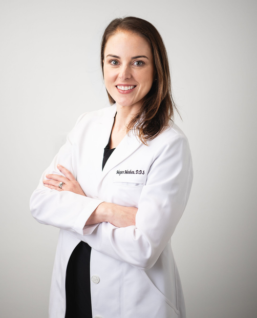 Dentist in a white coat smiling in a dental clinic, standing beside an exam chair.