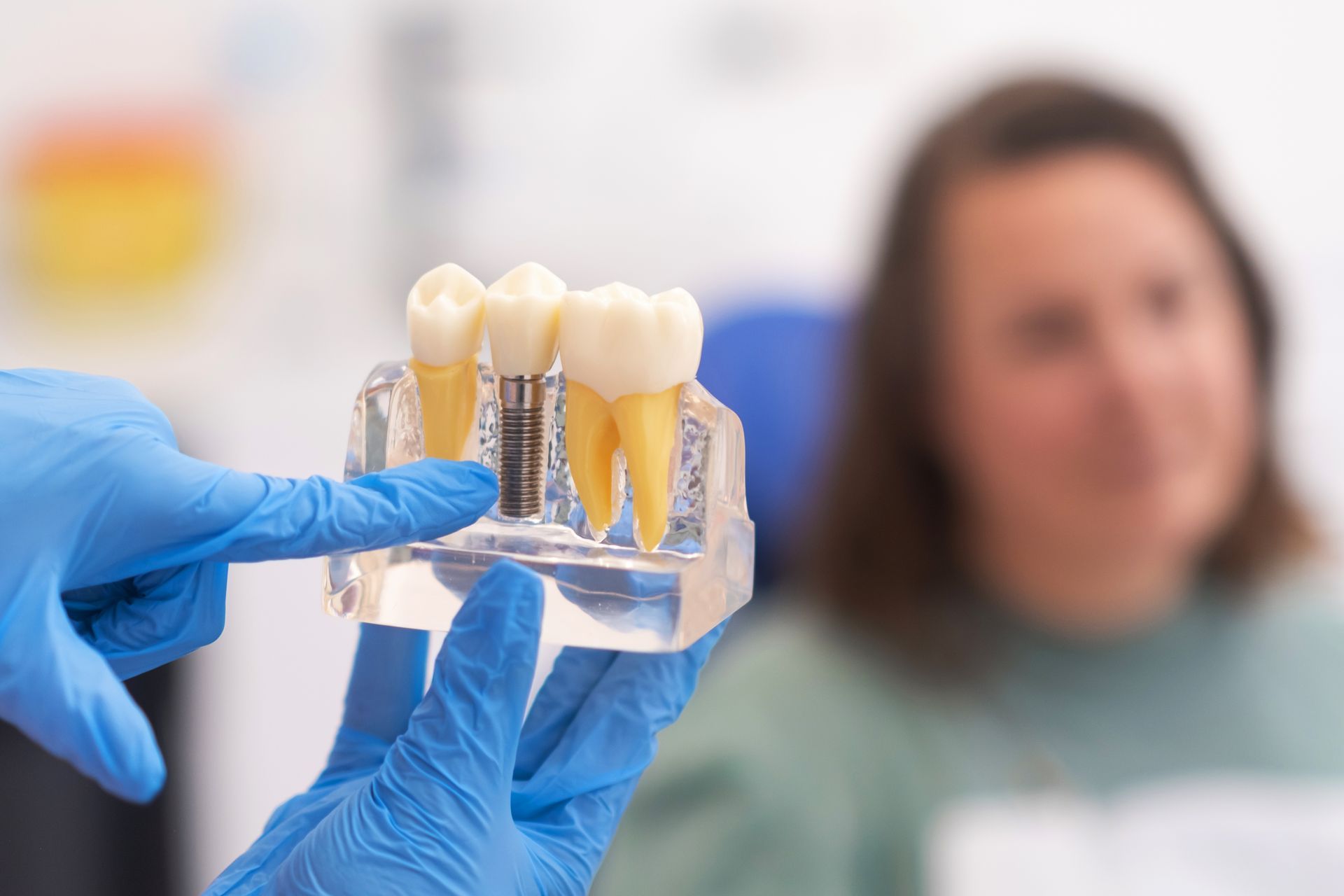 Gloved hands holding dental implant models with a blurred woman in a clinic background