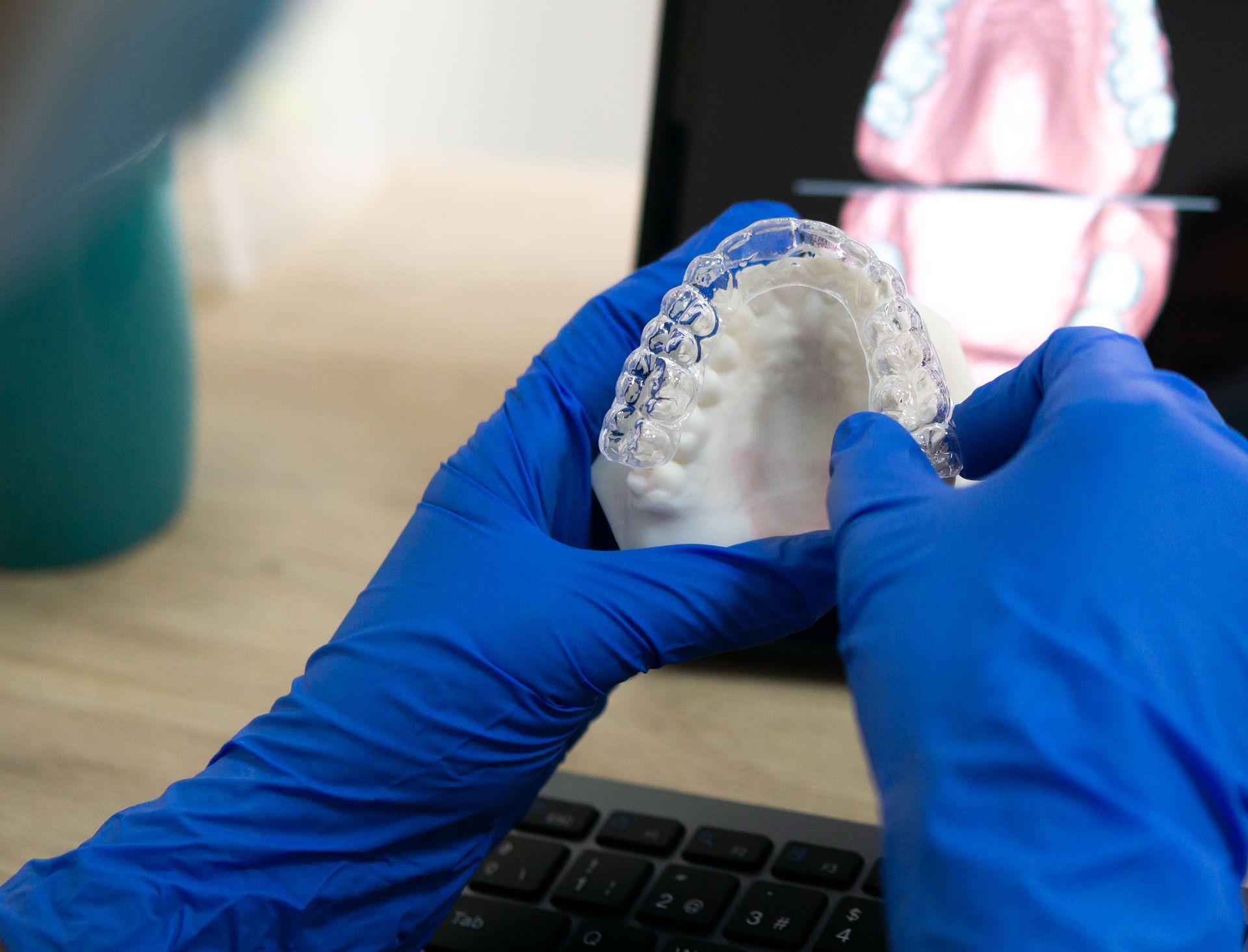 Blue-gloved hands holding a clear dental aligner in front of a laptop screen