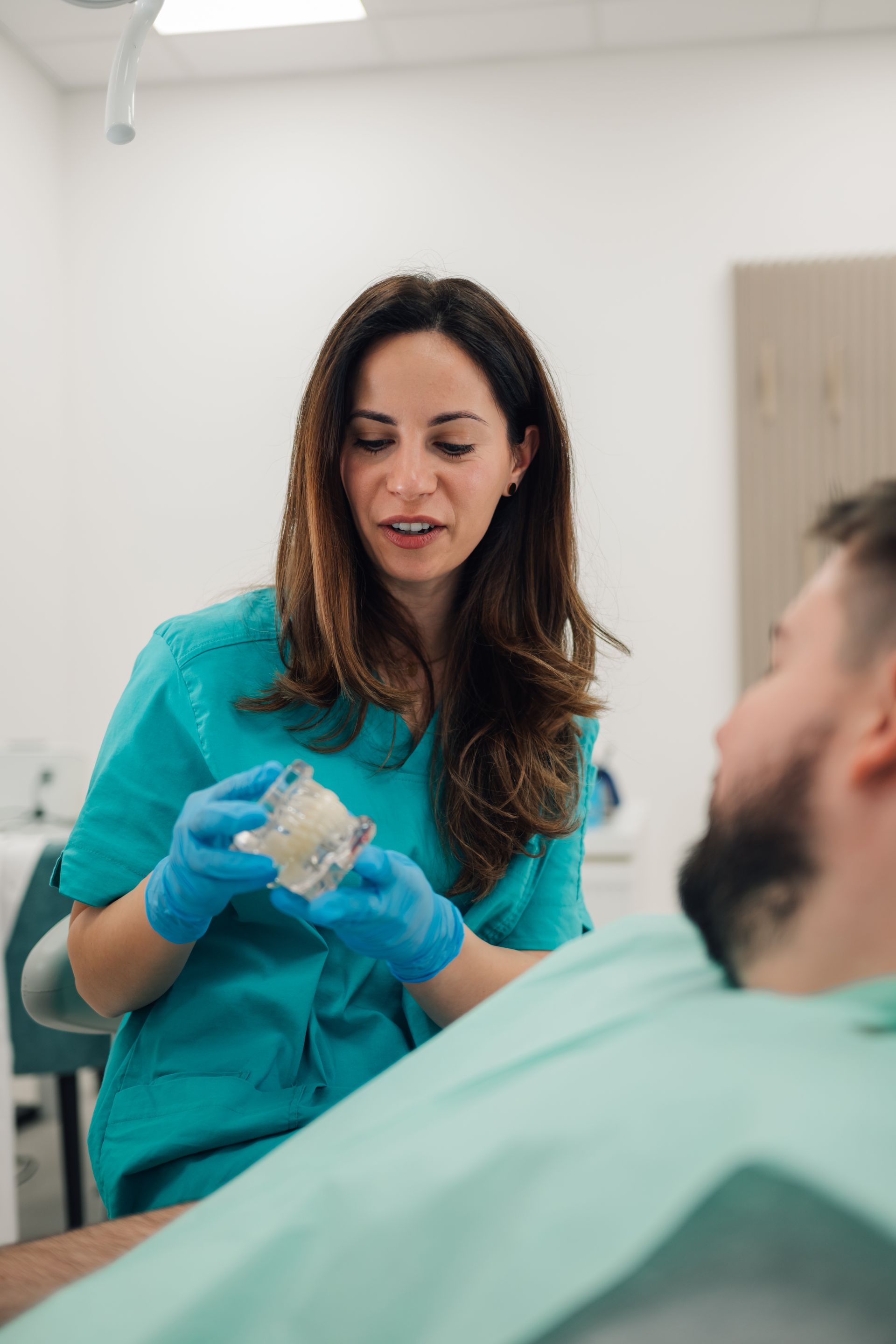 Dentist in teal scrubs and gloves showing a dental model to a seated patient in a clinic