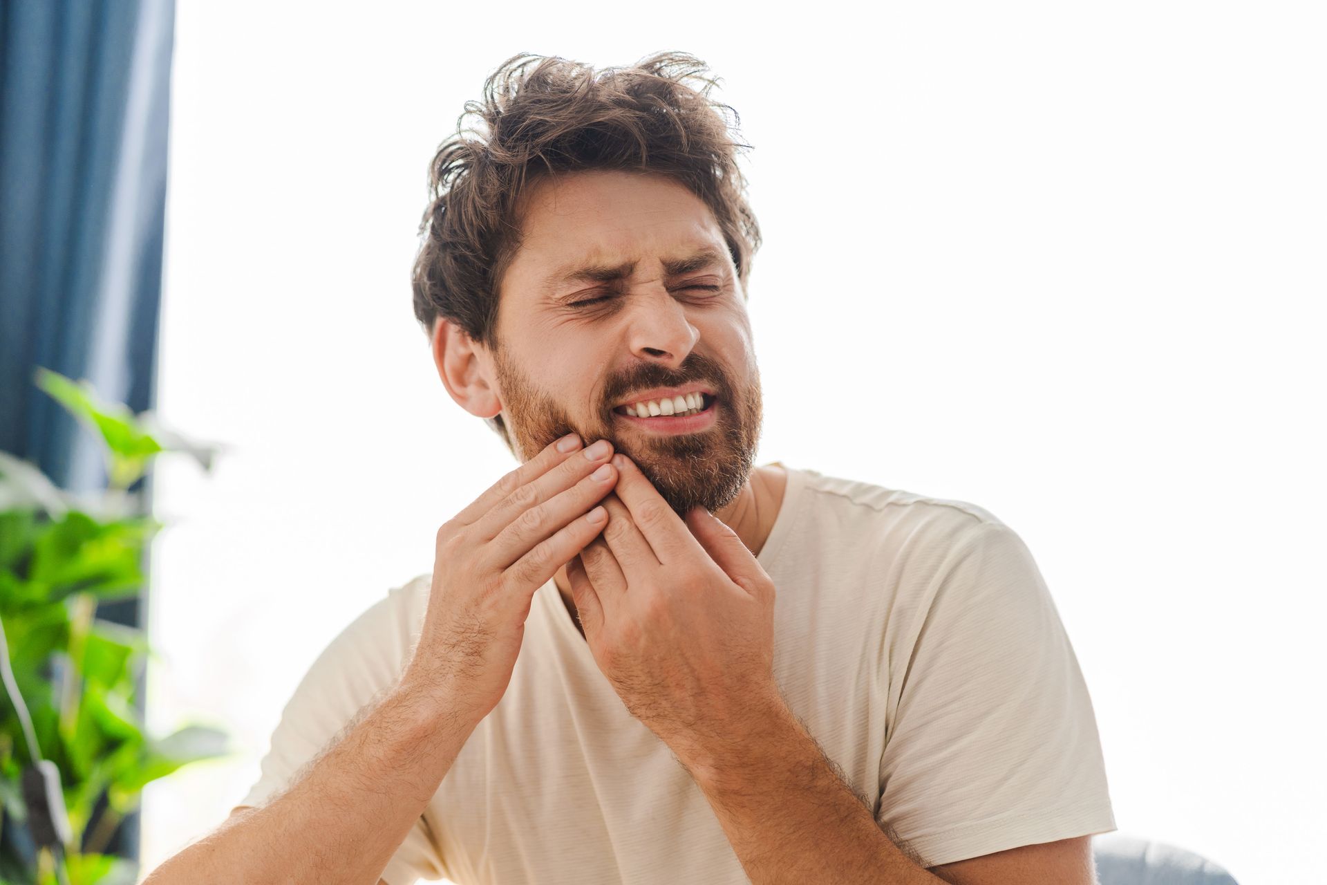 Man grimacing and holding his chin, appearing to have tooth pain in a bright room