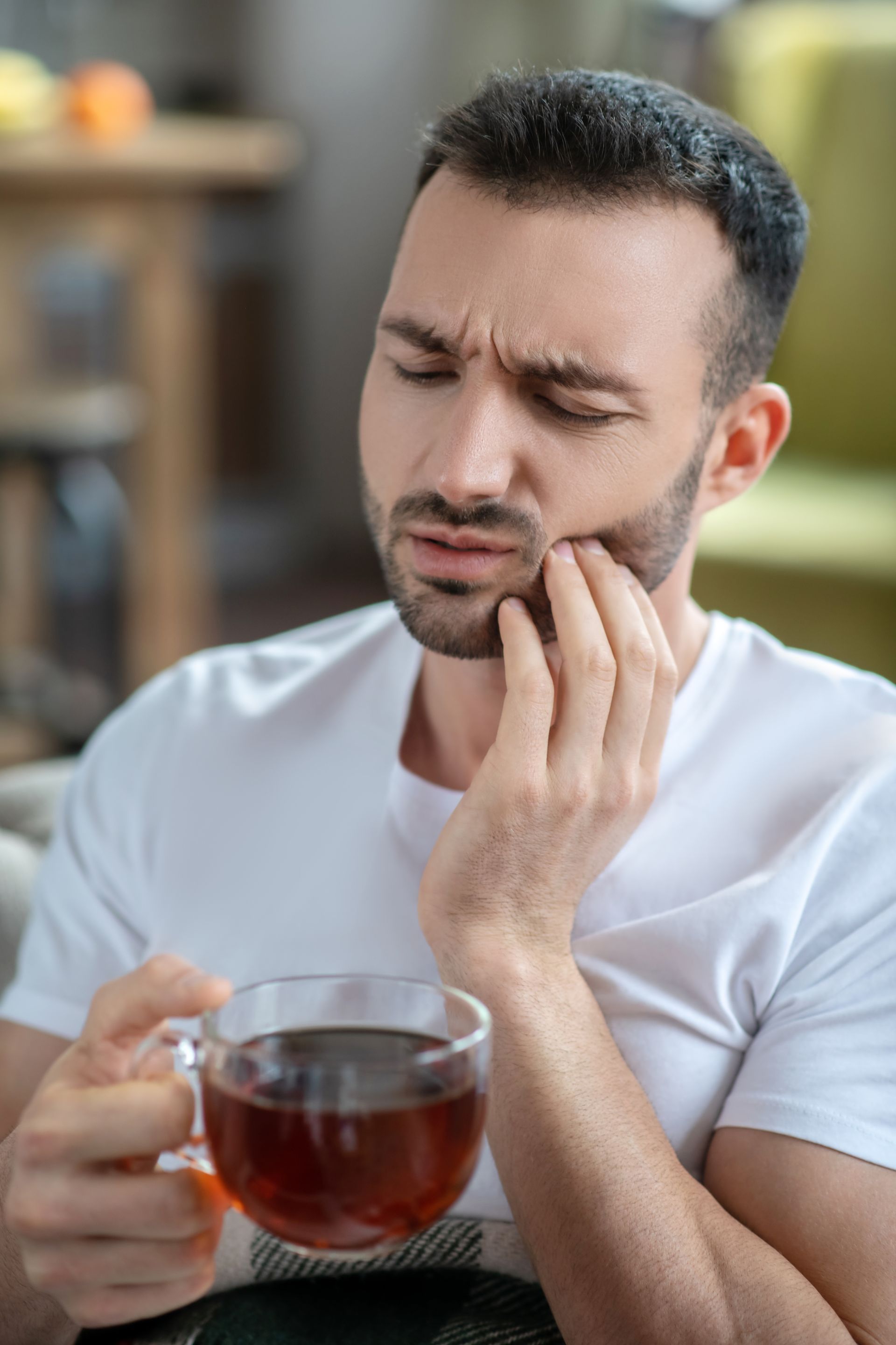 Man holding a mug of tea and touching his cheek, wincing in pain indoors