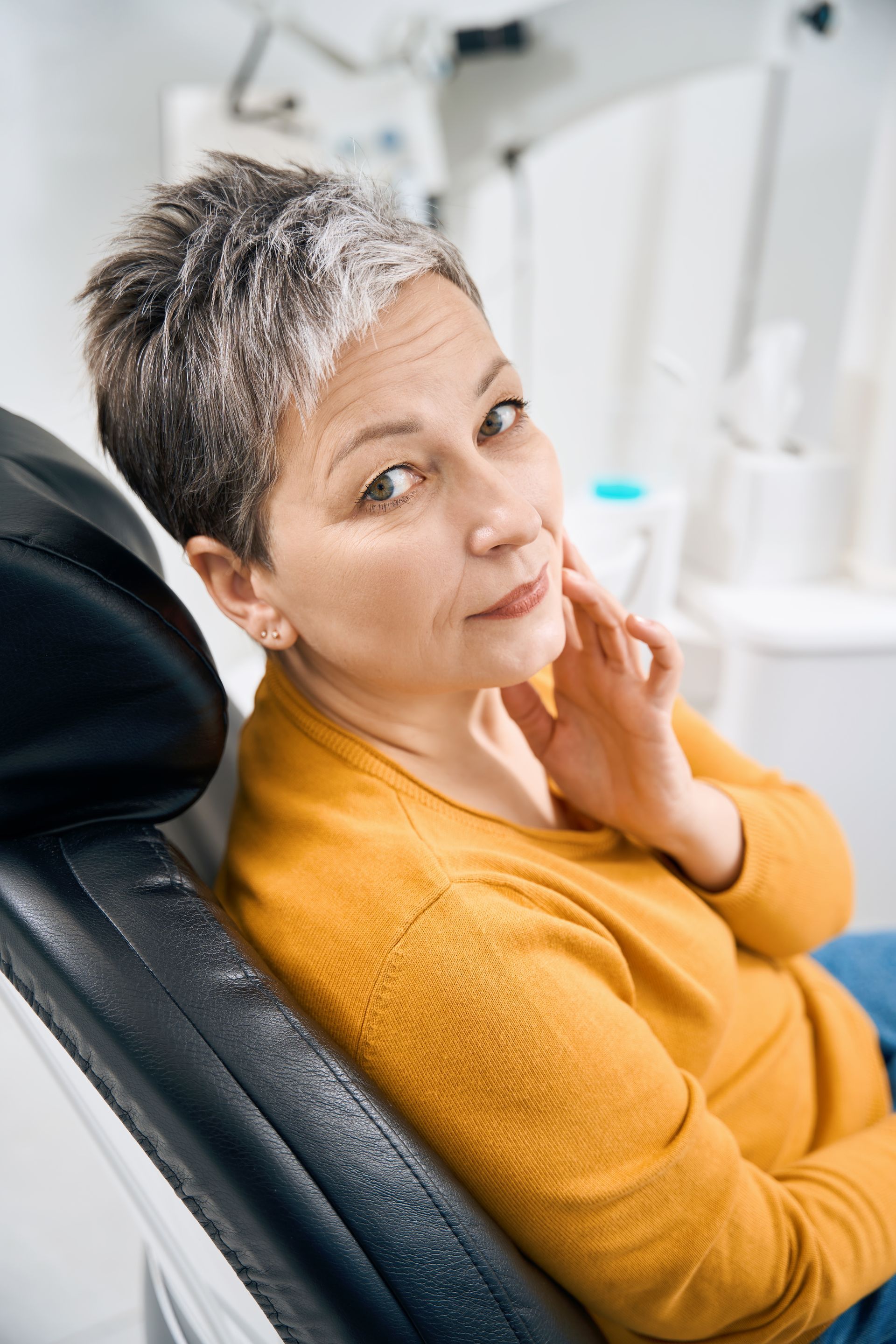 Person reclining in a dental chair, wearing a mustard sweater and looking at the camera with a hand on their cheek