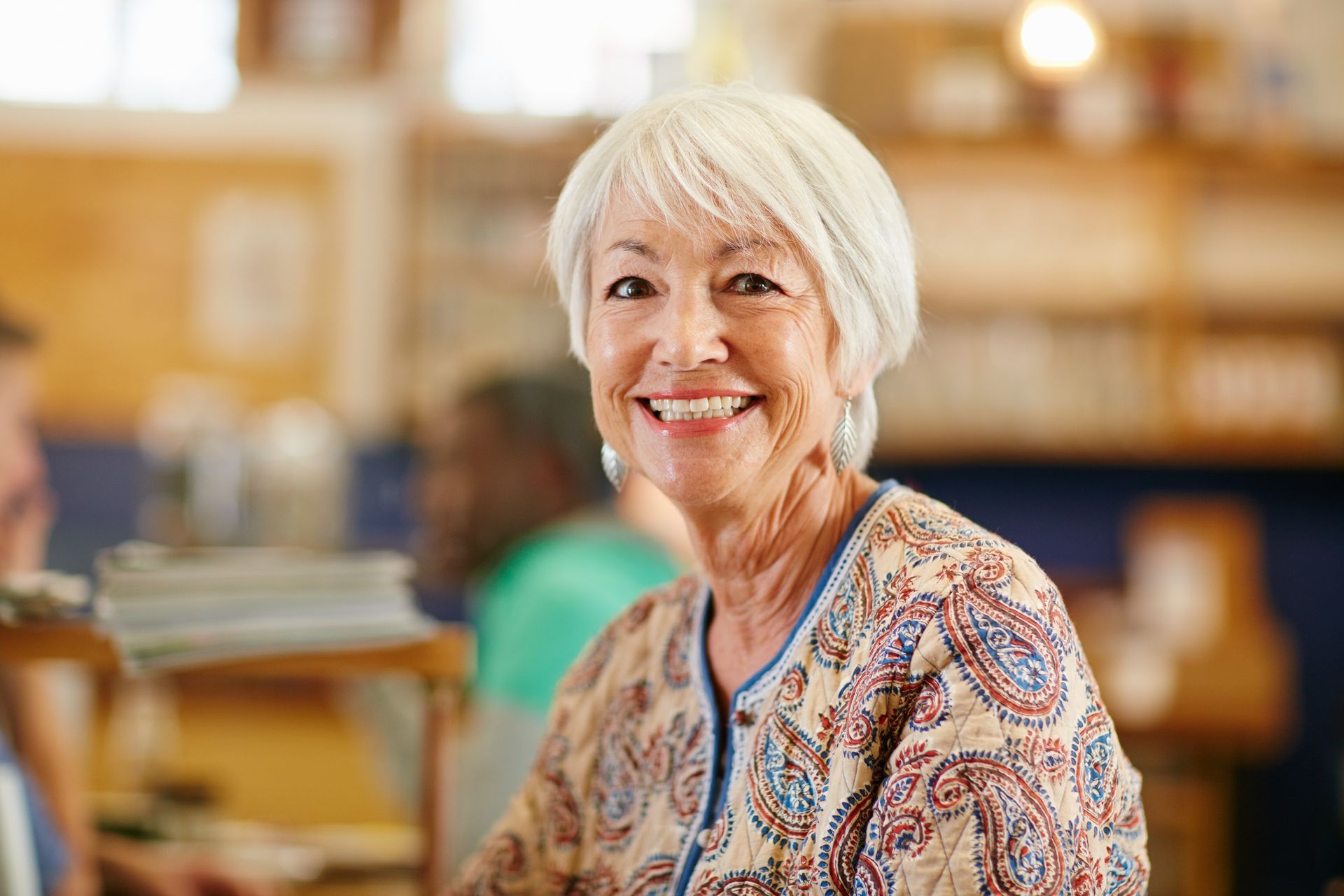 Smiling person in a patterned blouse seated indoors, with a softly blurred background.