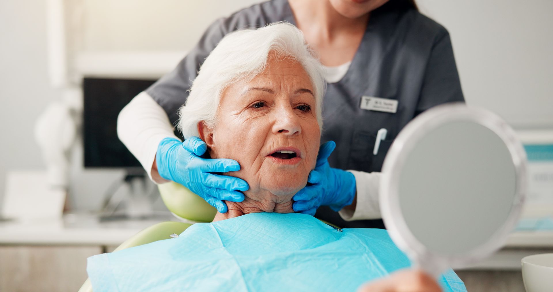Dental hygienist treating a seated patient wearing a bib in a clinic, with blue gloves and a mirror nearby