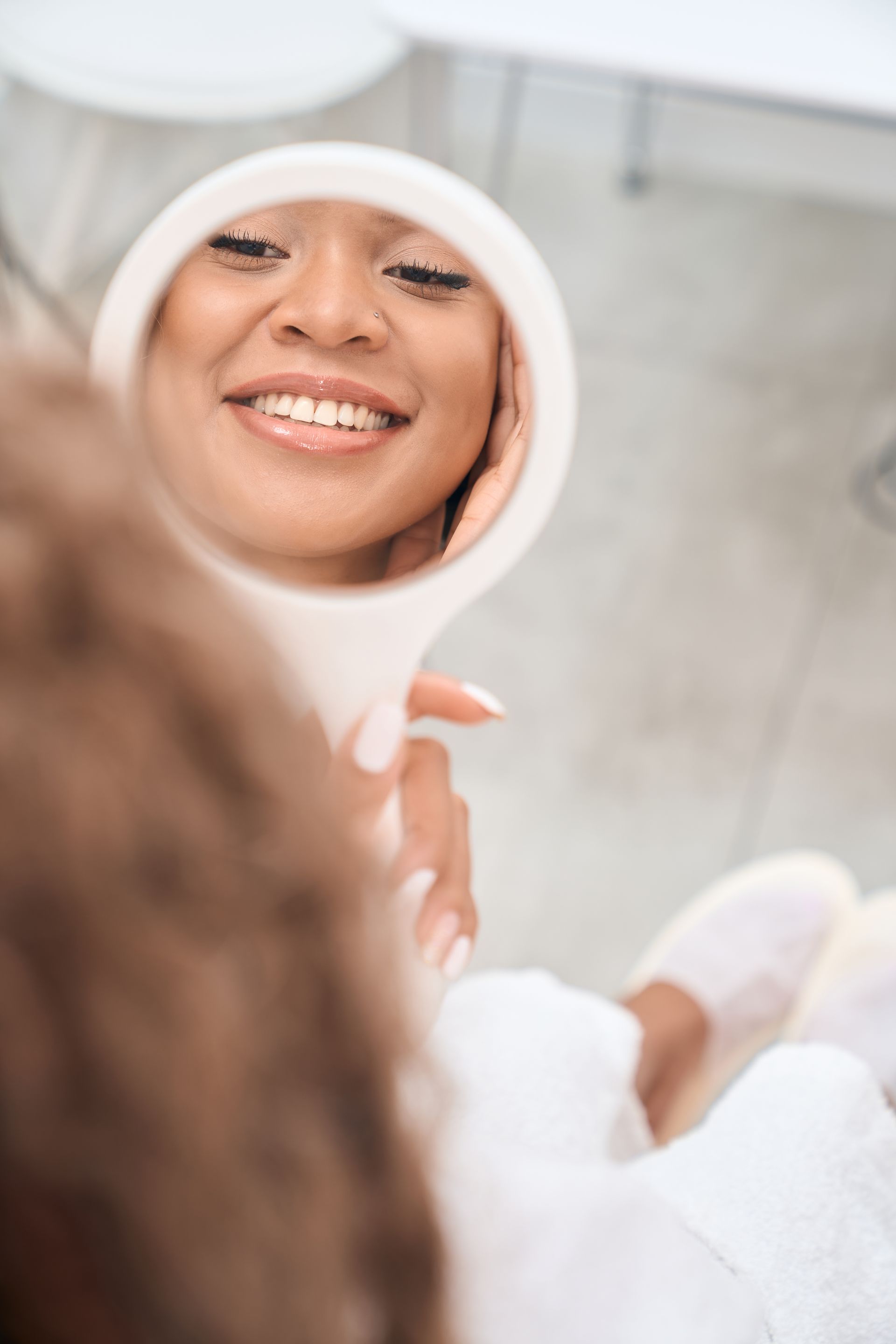 Person smiling into a handheld mirror in a bright bathroom