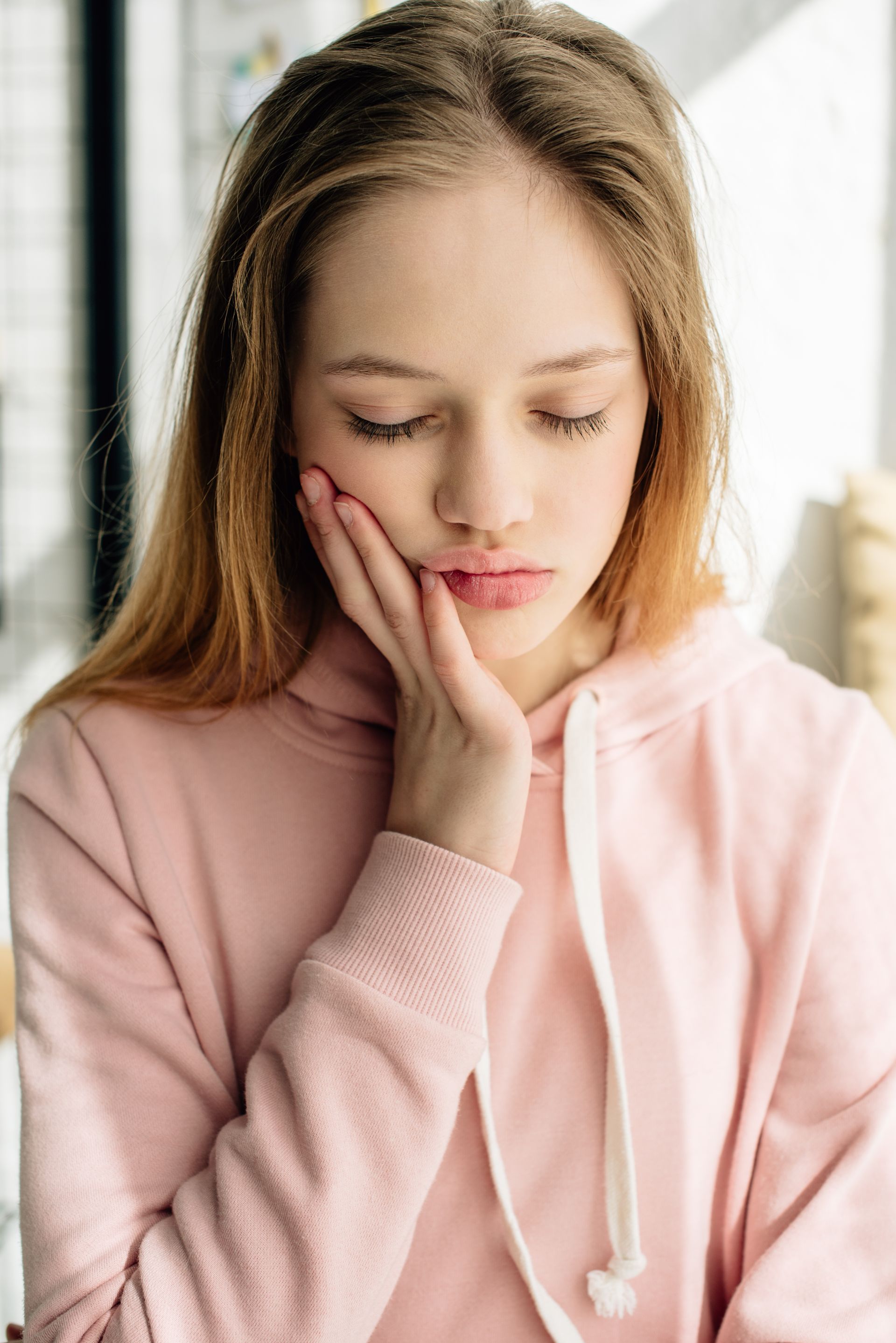 Young woman in a pink hoodie resting her face on her hand, looking down thoughtfully by a window