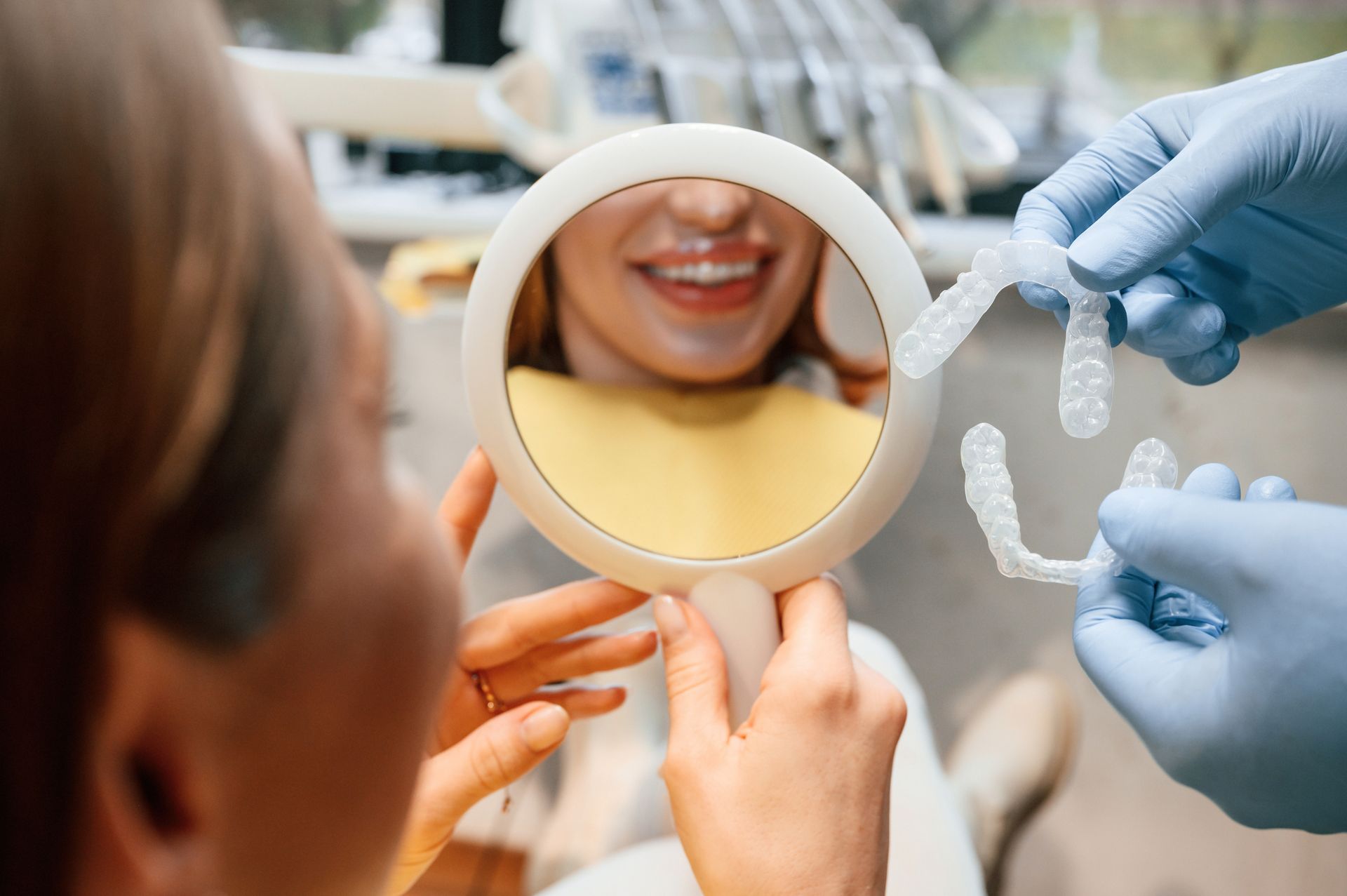 Hands fitting a dental retainer in front of a smiling person holding a mirror.