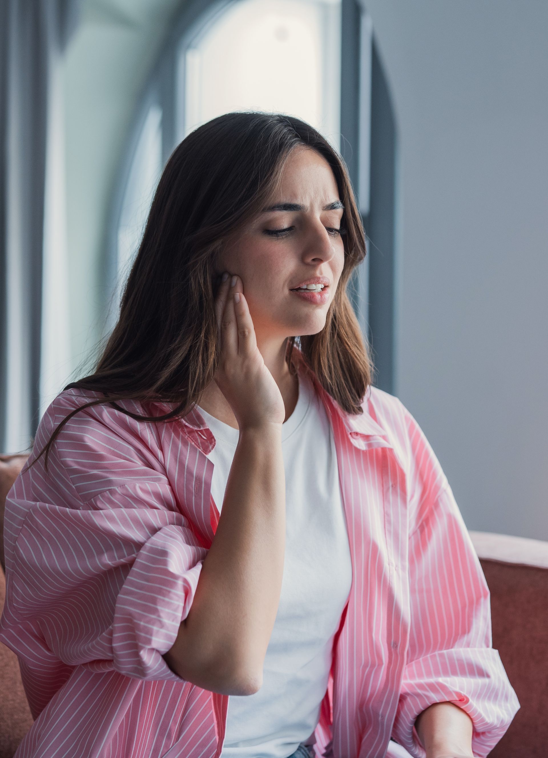 Woman in a pink striped shirt touching her cheek by a window, looking pensive.