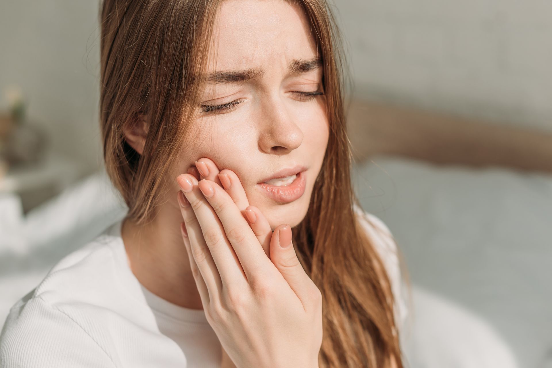 Woman holding her cheek with eyes closed, appearing to have tooth pain on a bed