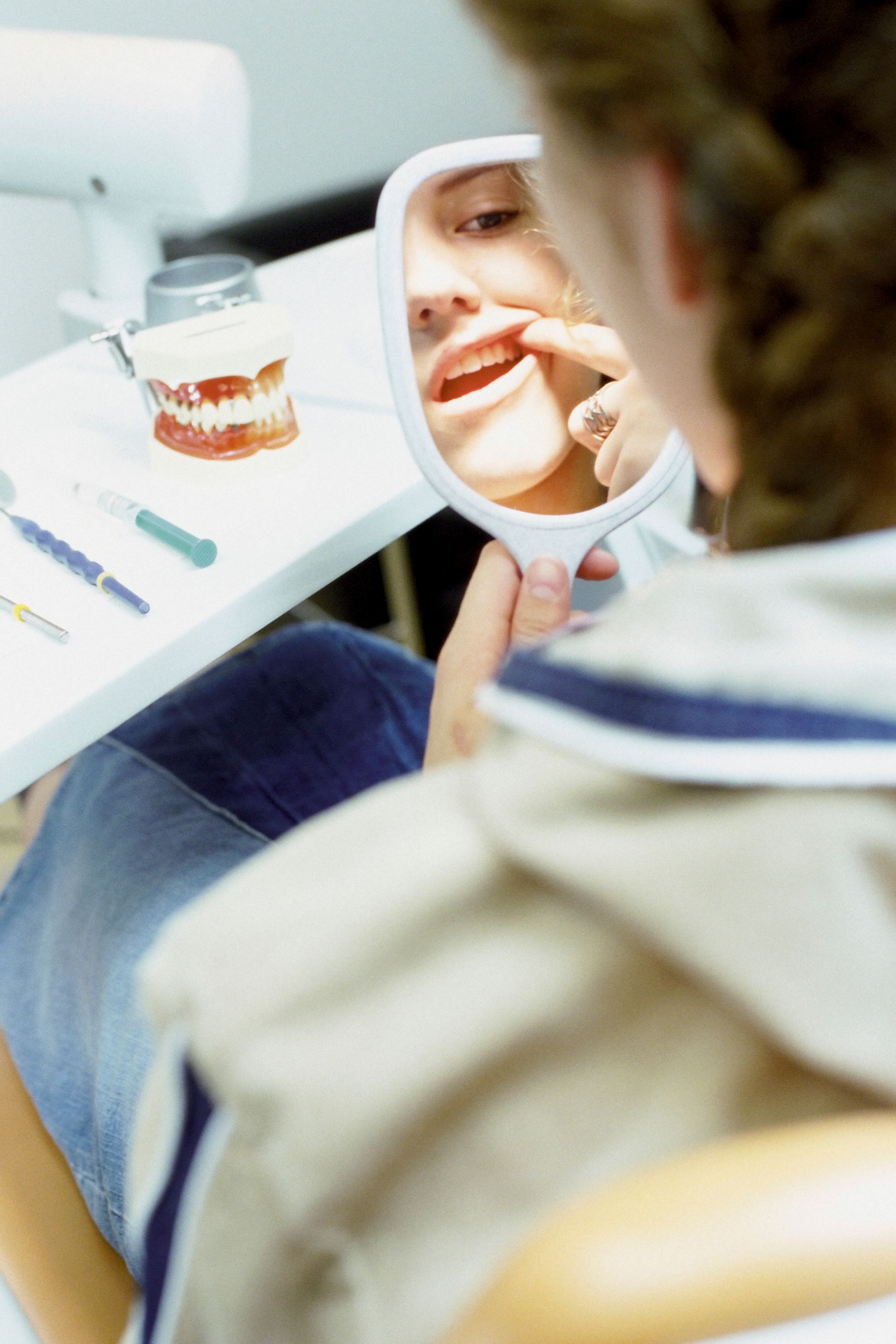 Person looking into a hand mirror while holding a toothbrush, with a glass and toothbrushes on a table nearby.