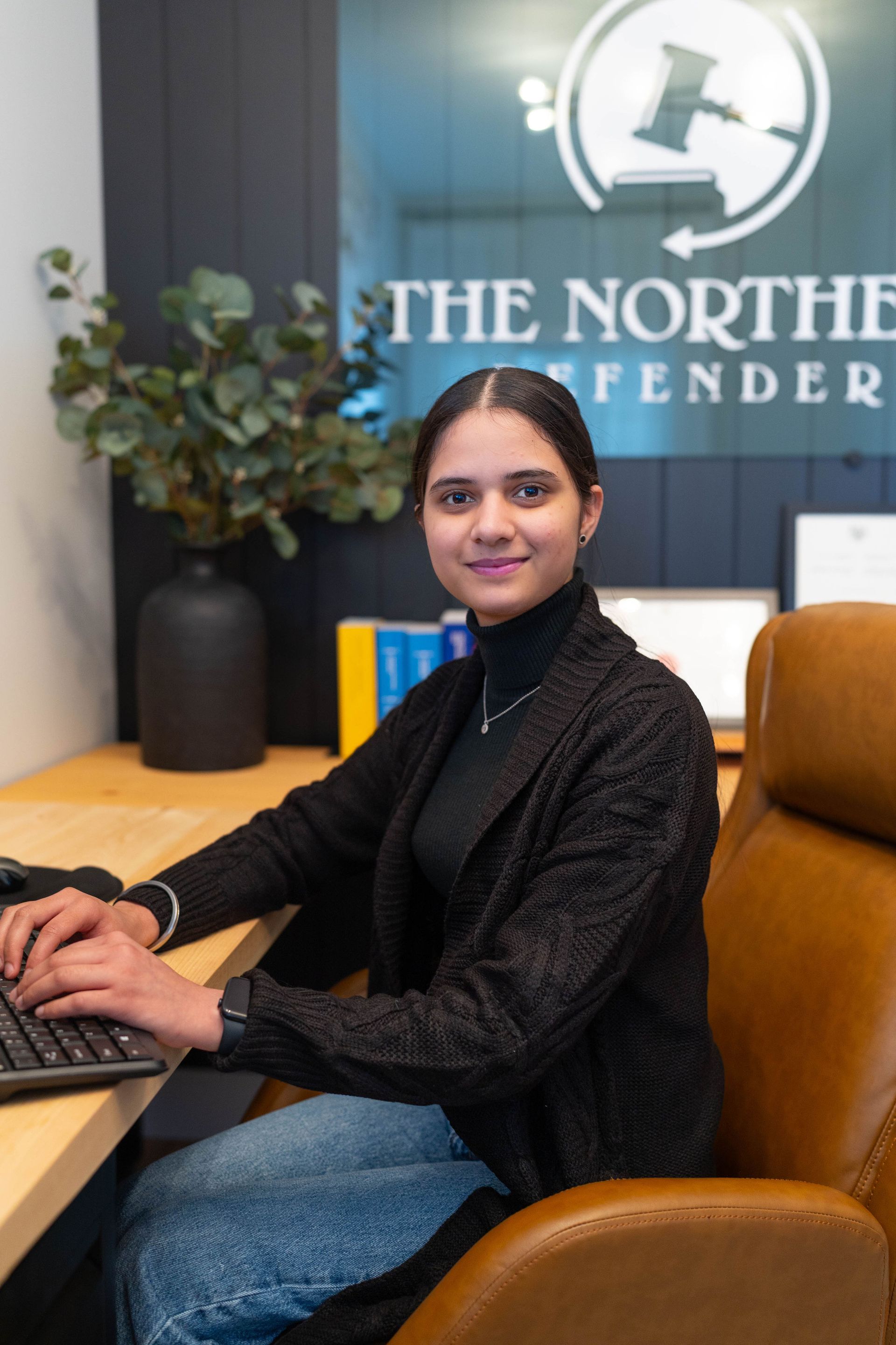 Woman seated at a desk typing in an office with “The Northern Defender” sign in the background