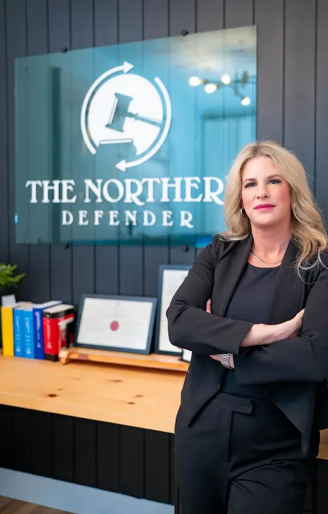 Woman standing with arms crossed beside a “The Northern Defender” office sign and desk
